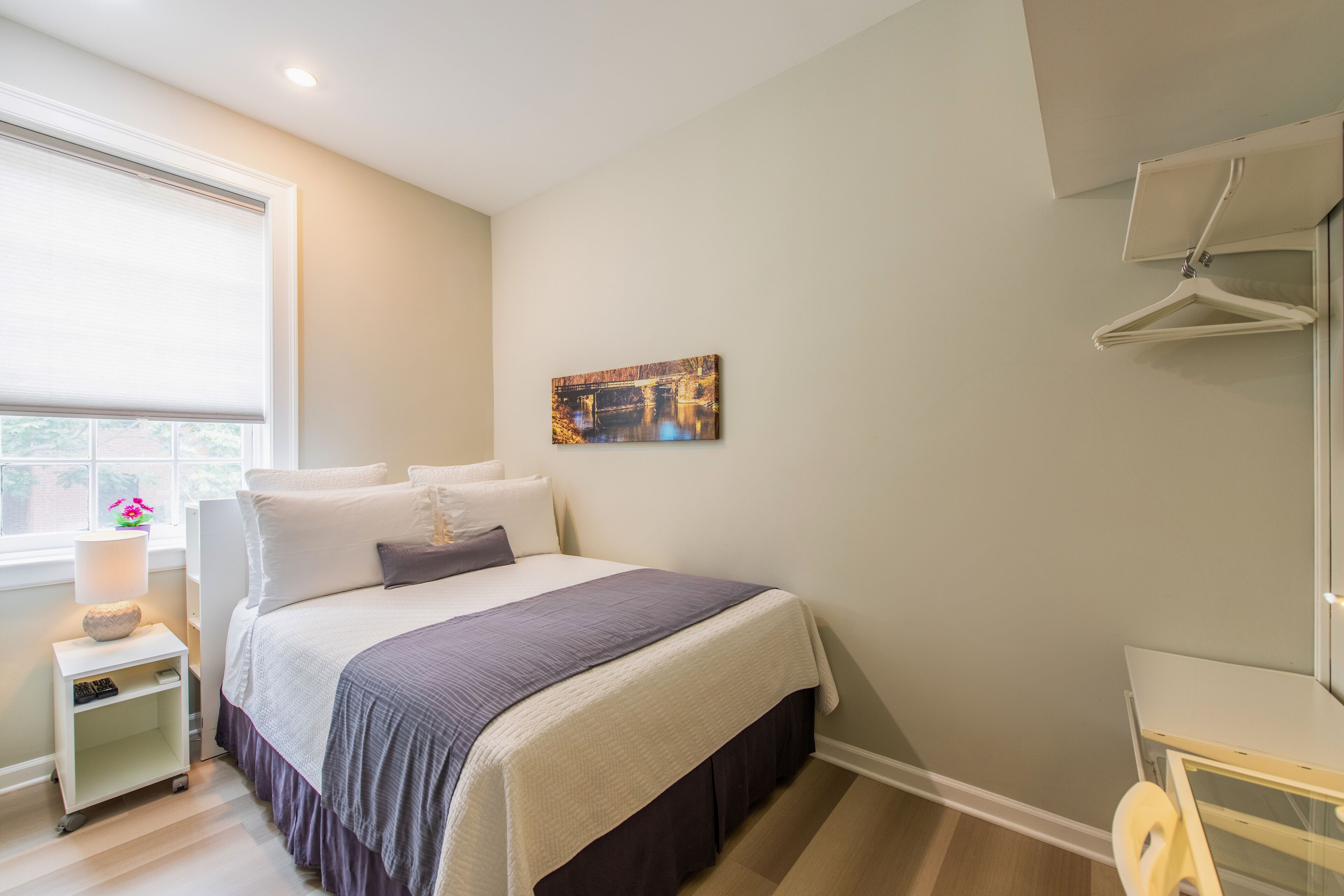 A bedroom with a bed, a nightstand, a lamp, a painting, and a window.