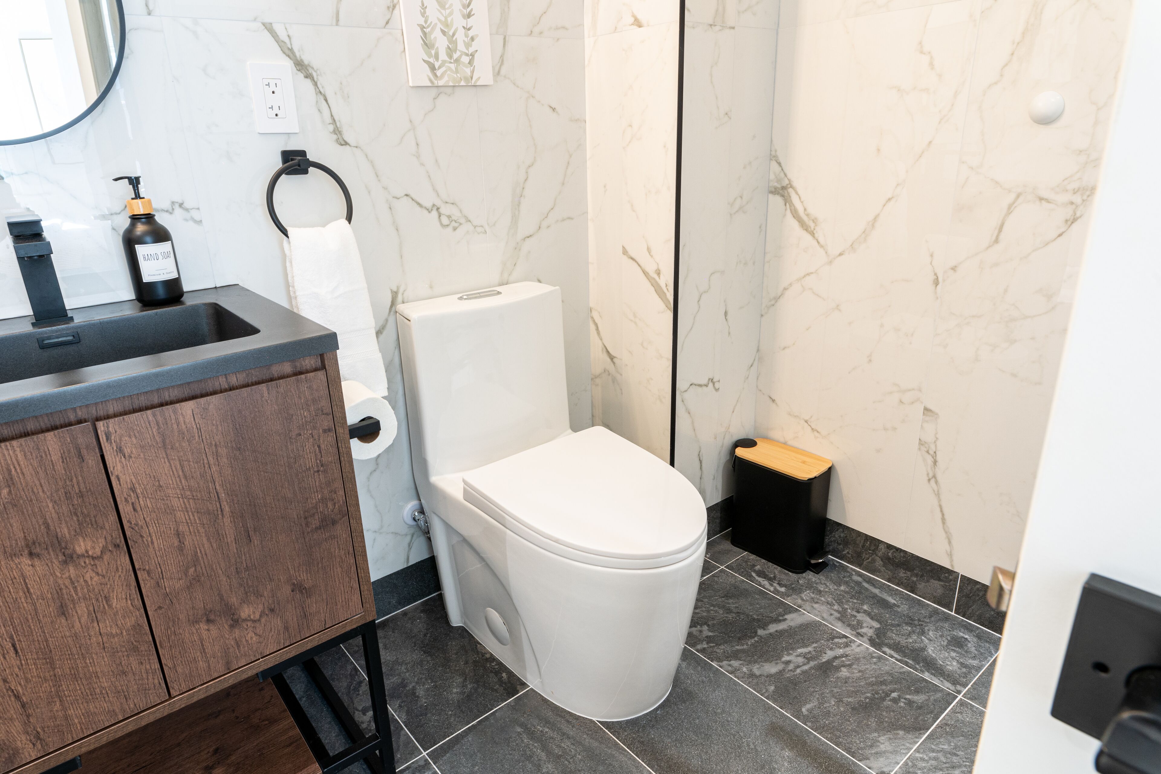 Un baño moderno con pared de mármol, tocador de madera, inodoro blanco y cubo de basura negro.