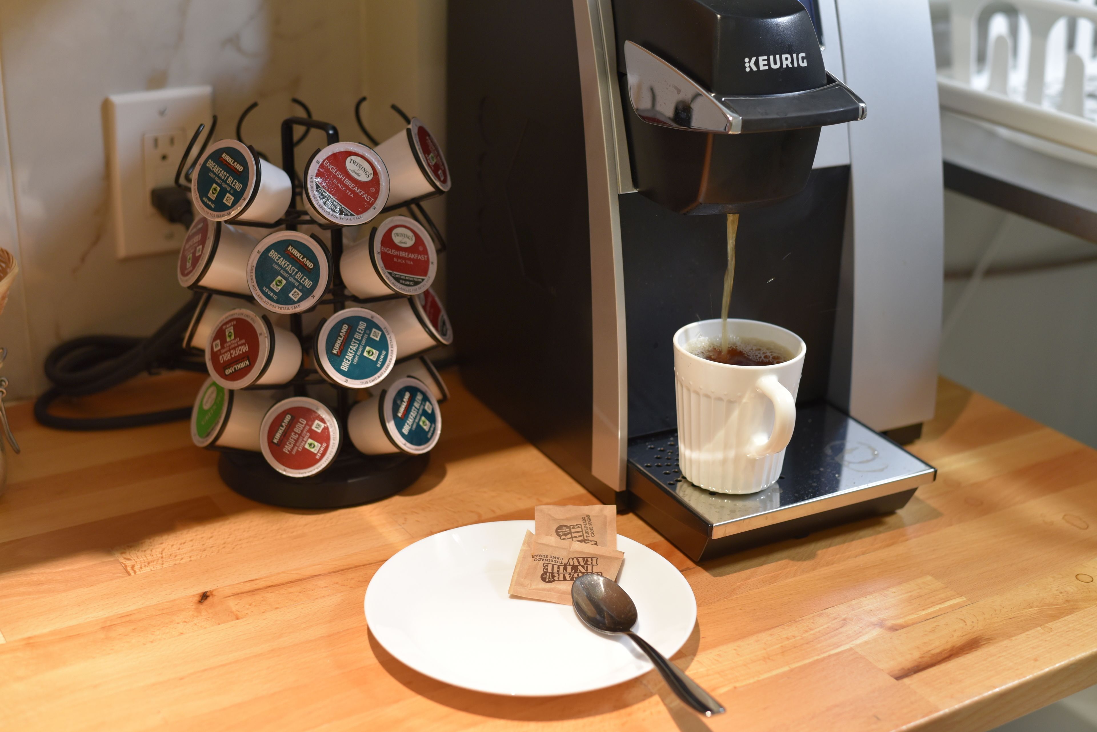 Une machine à café qui verse du café dans une tasse, un support pour dosettes de café et une cuillère sur une assiette blanche.