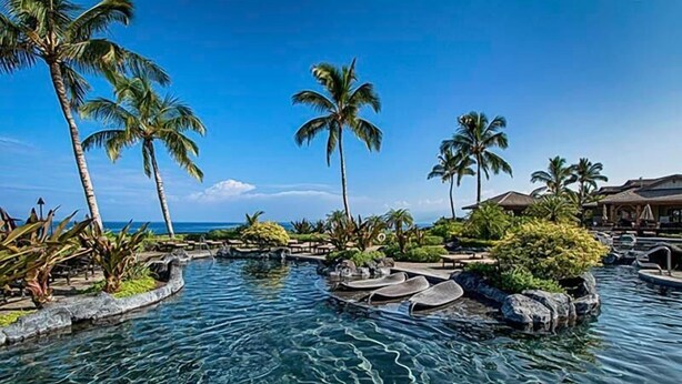 Zwembad van een resort met helder water, palmbomen en uitzicht op de oceaan.
