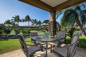 A covered outdoor dining area with a table and chairs, overlooking a well-maintained garden and a golf course. - Hali i Kai 18 H (Waikoloa)