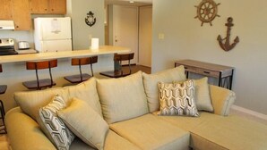 A living room with a beige sectional sofa, wooden chairs, a small kitchen area with a refrigerator, and nautical decor on the walls. - Lake Arrowhead Fiesta (Myrtle Beach)