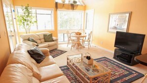 A living room with a beige sofa, a glass coffee table, a TV, and a dining area with chairs and a table. - Lake Arrowhead Den (Myrtle Beach)