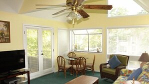 Ein Wohnzimmer mit Deckenventilator, Fernseher auf einem Schrank, einem Esstisch mit Stühlen und einer floral gemusterten Couch.