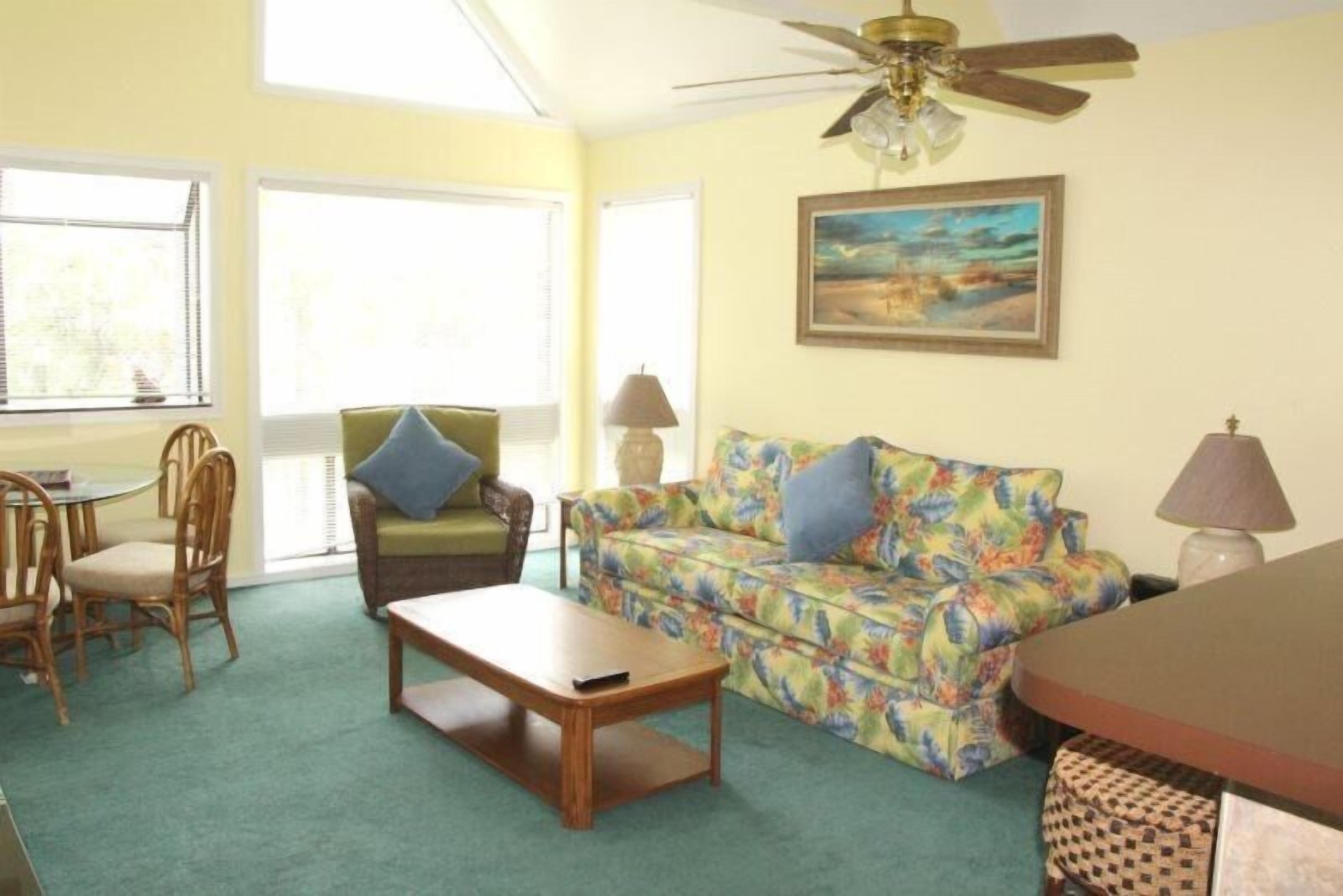 A living room with a green carpet, a wooden coffee table, a floral sofa, a dining table with chairs, and a ceiling fan.