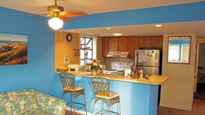 A kitchen with a blue accent wall, wooden cabinets, a central island with bar stools, a ceiling fan, and a refrigerator. - Lake Arrowhead Casa (Myrtle Beach)