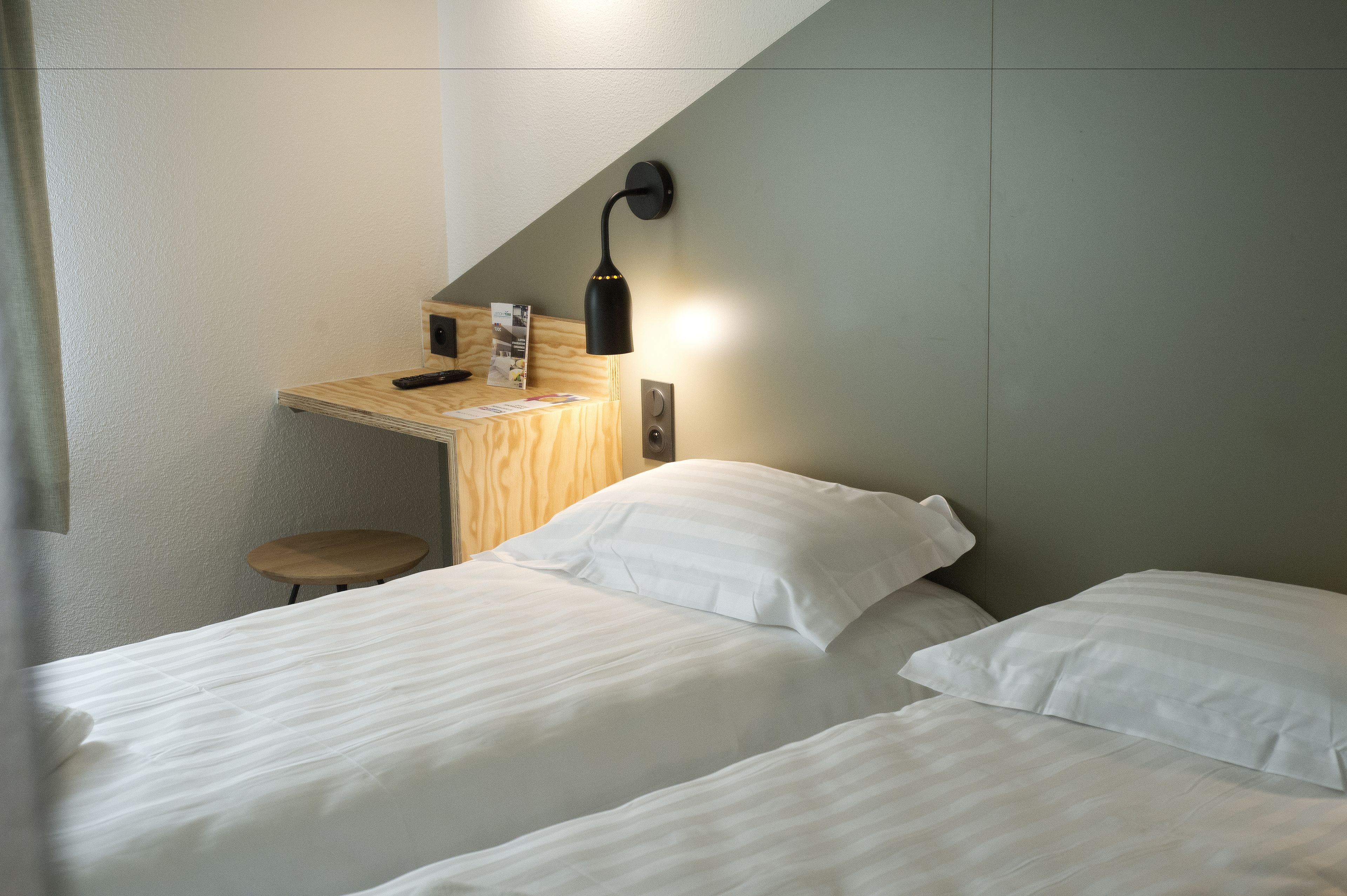 A hotel room with two beds, a desk, a chair, and a wall-mounted lamp.
