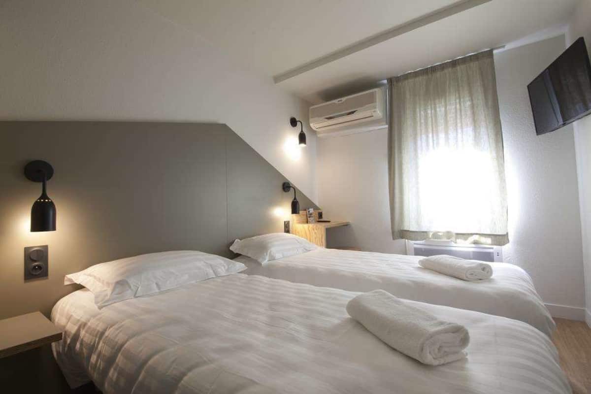 A hotel room with two beds, a bedside table, a wall-mounted air conditioner, a TV, and a window with curtains.