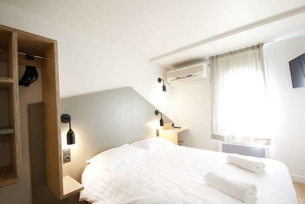A hotel room with a bed, a wooden wardrobe, a bedside table with a lamp, an air conditioning unit, and a window with curtains.