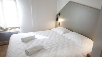 A neatly made bed with white linens and towels, a modern headboard, and a wall-mounted light fixture.