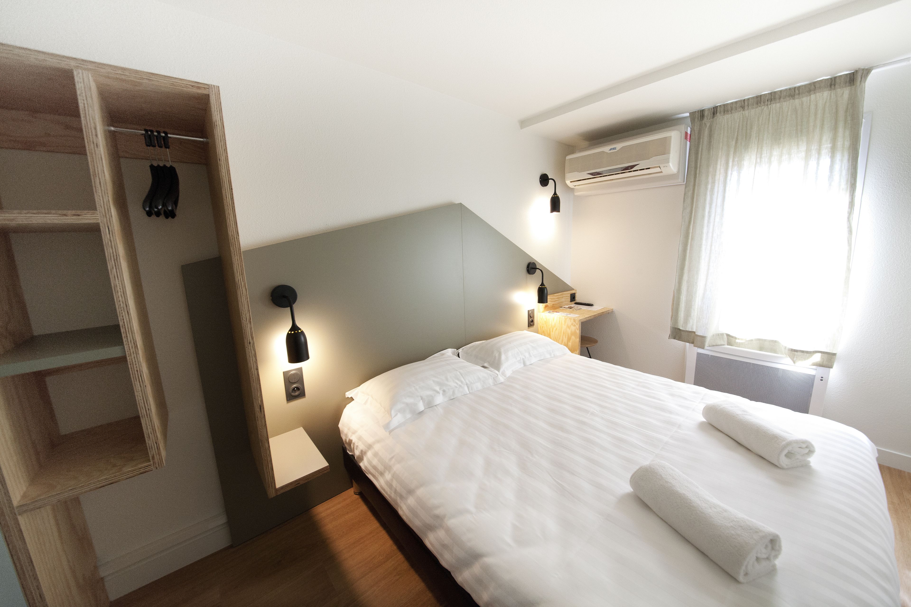 A neatly made bed with white linens and two rolled towels, a wooden wardrobe with hanging items, a bedside table with a lamp, and a window with curtains.