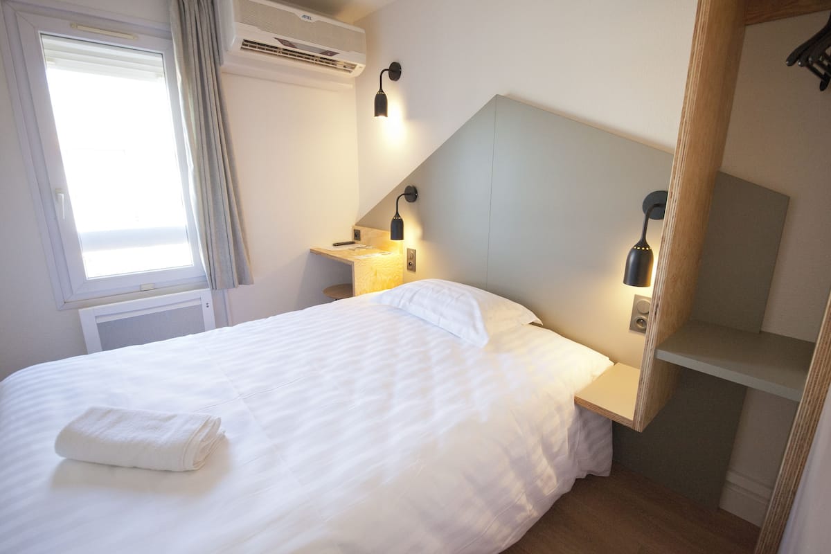 A neatly made bed with a folded towel, a bedside table with a lamp, and a small shelf.