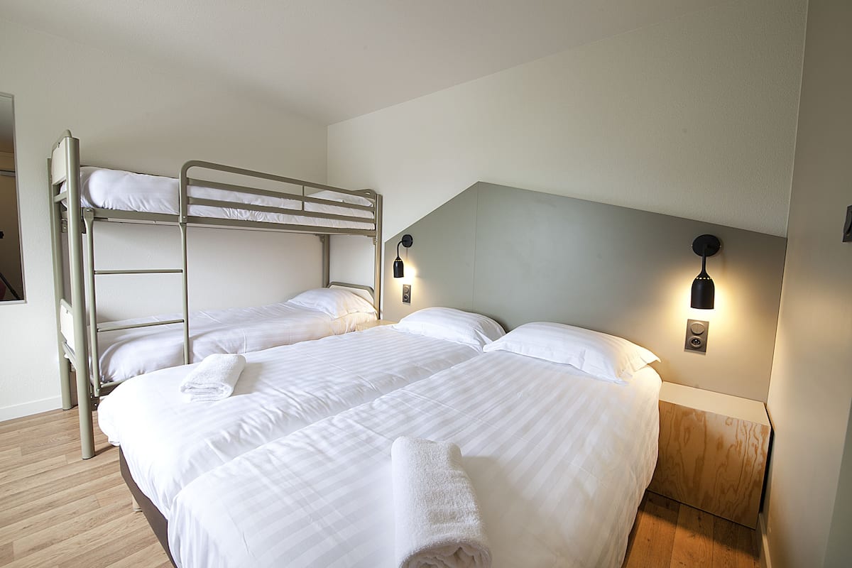 A bunk bed room with two beds, a wooden nightstand with a lamp, and a wooden floor.