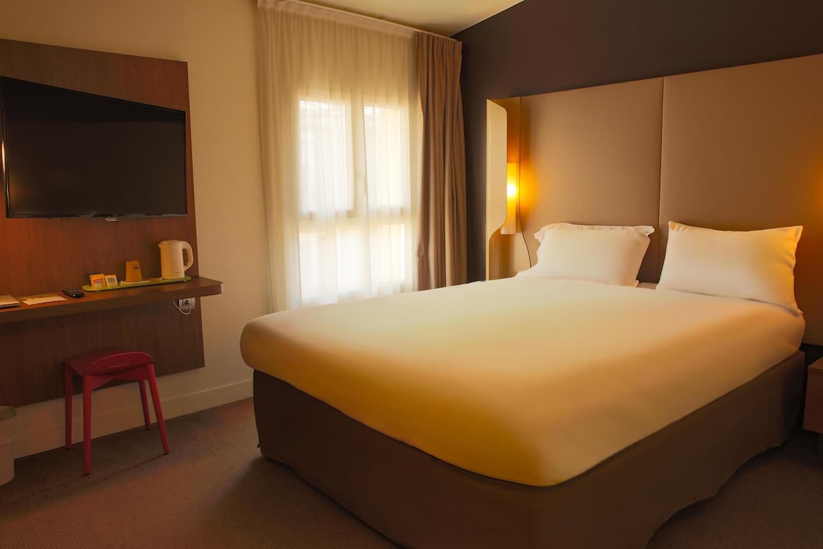 Une chambre d’hôtel dotée d’un grand lit, d’un téléviseur à écran plat fixé au mur, d’une table de chevet avec une lampe et d’une fenêtre ornée de rideaux légers.