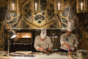Dos cocineros preparando comida en una cocina con paredes de mármol e iluminación colgante.