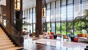 A spacious lobby with a marble floor, a staircase, and large windows offering a view of greenery outside.