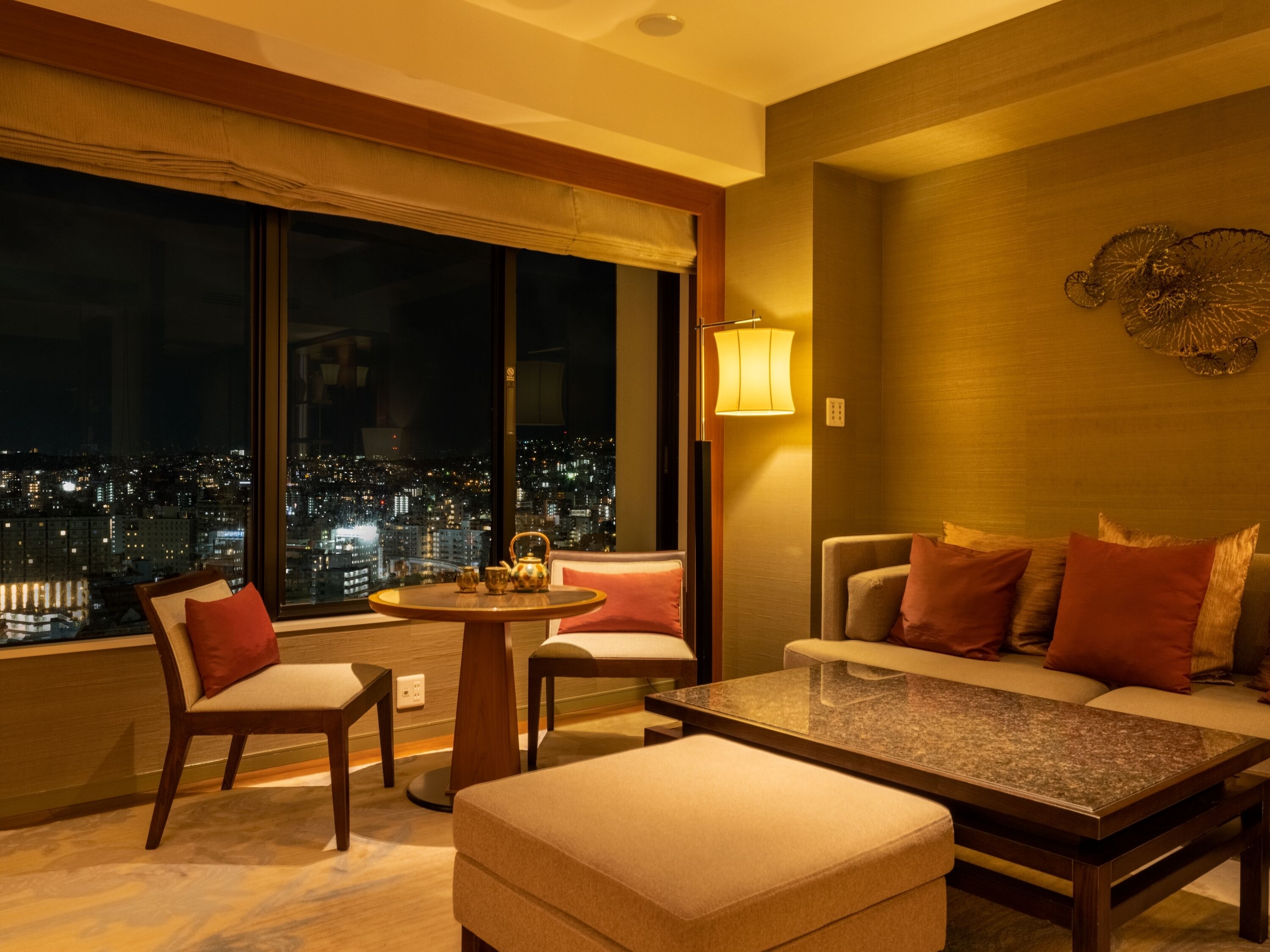 A modern hotel room with a city view, a round table, a sofa, and two armchairs.