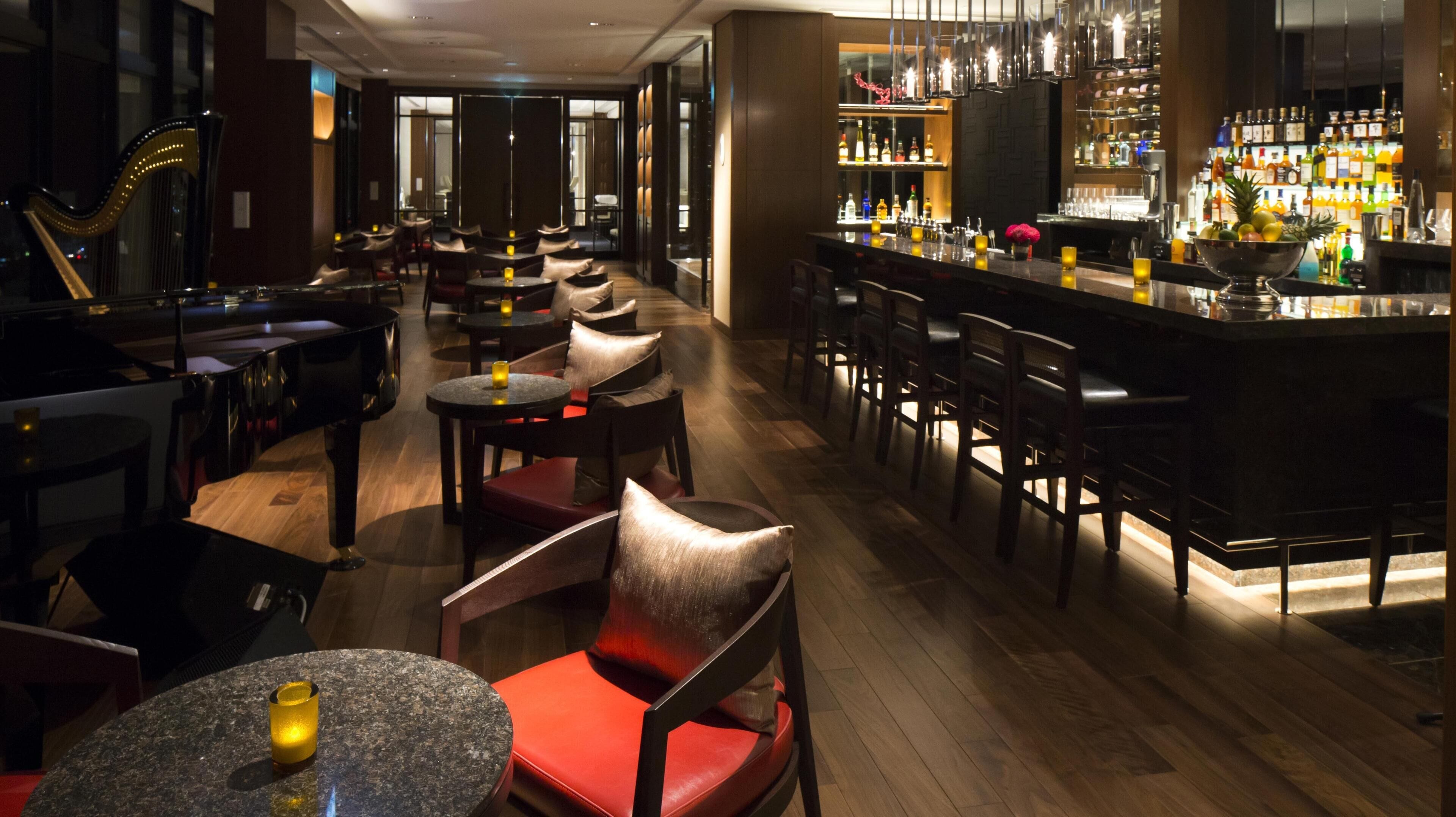 A bar area with multiple seating options, including round tables and bar stools. There is a piano in the foreground, and the bar counter is stocked with various bottles.