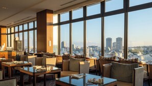 A modern hotel lobby with large windows offering a city view, comfortable seating areas, and a bar with a cityscape visible through the windows.