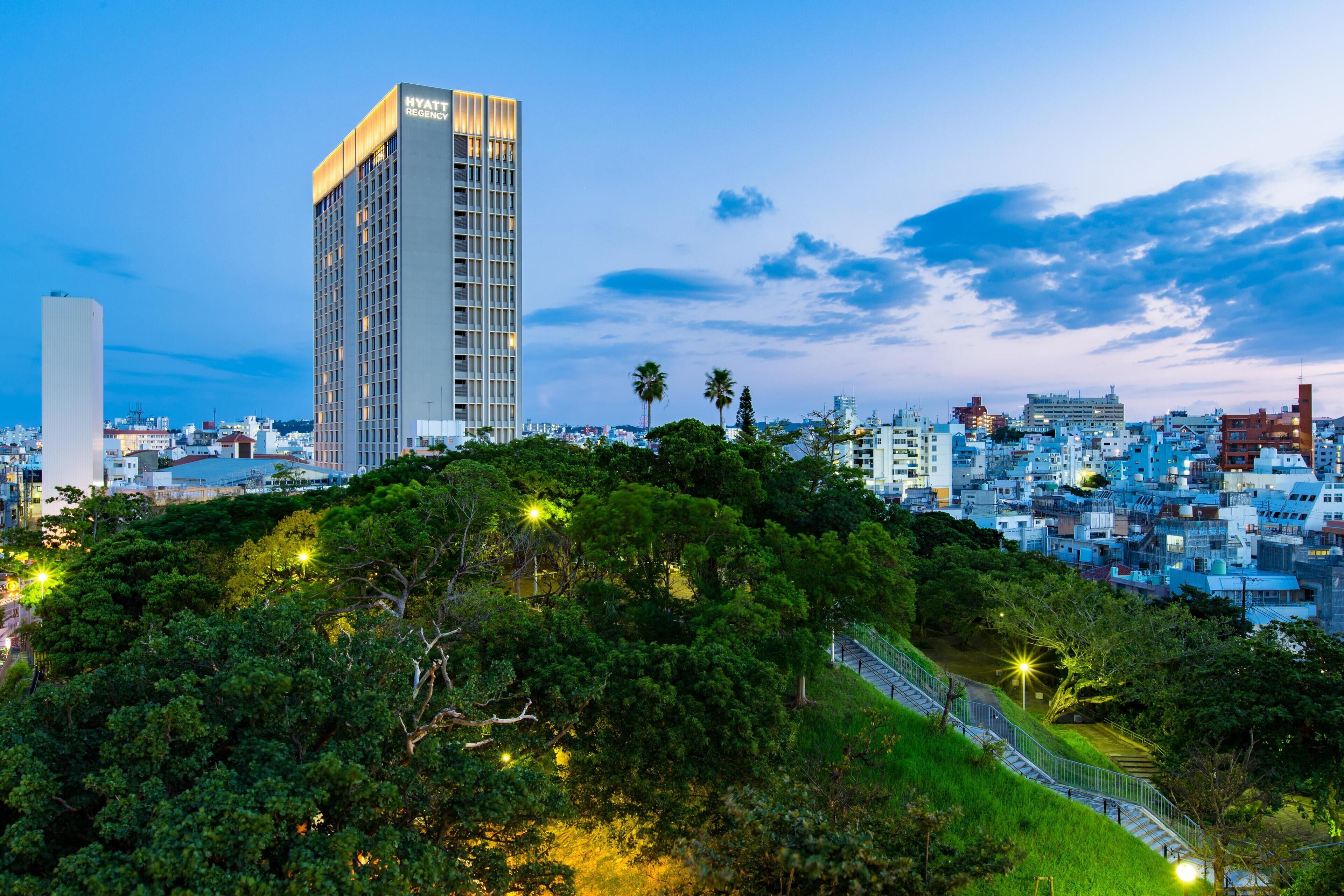 A high-rise hotel with a modern design, surrounded by lush greenery and a cityscape in the background.