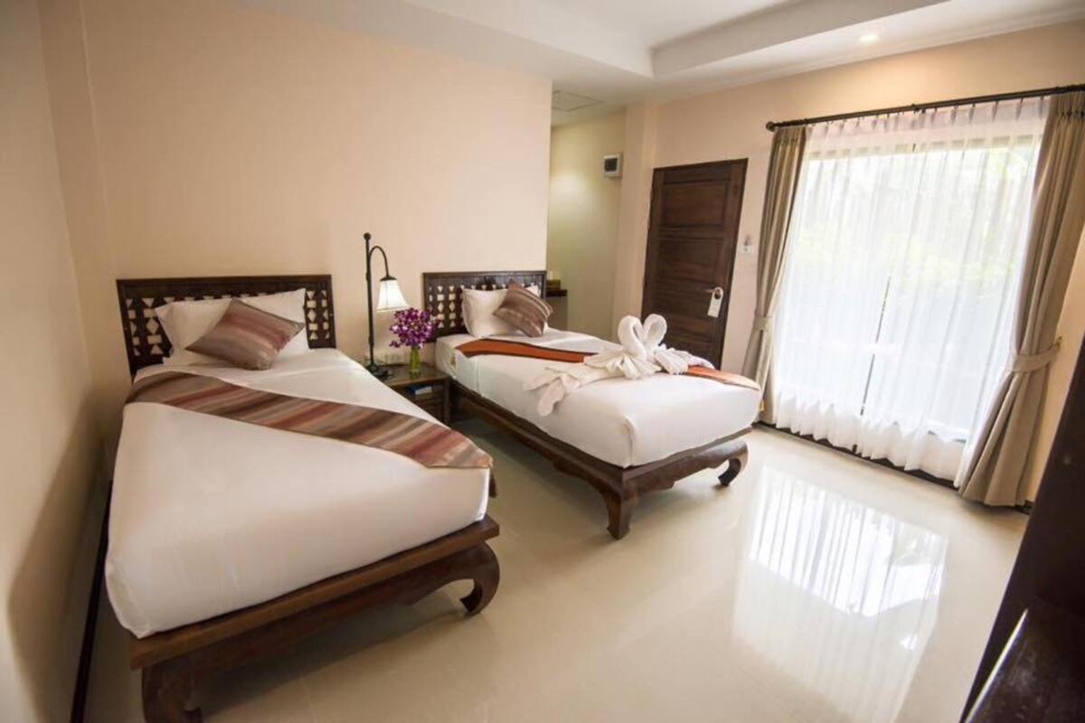 Habitación de hotel con dos camas, una mesita de noche con lámpara y un ventanal con cortinas.