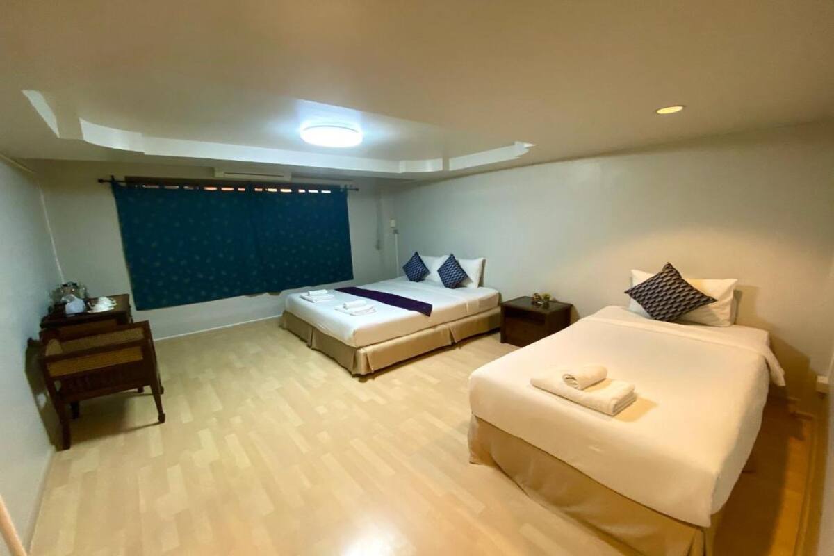 Una habitación de hotel con una cama grande, una cama más pequeña, un banco de madera, una mesita de noche y una ventana con cortinas.
