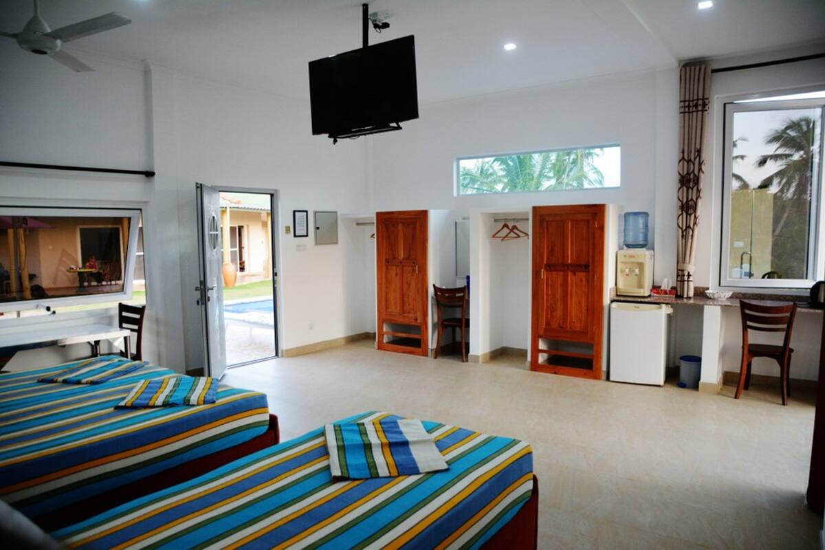 A hotel room with two beds, a TV, a desk with a computer, a window with a view of palm trees, and a door leading outside.