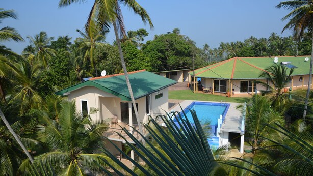 A resort with a swimming pool, palm trees, and a green-roofed building. - Hilltop Boosa (Boossa)