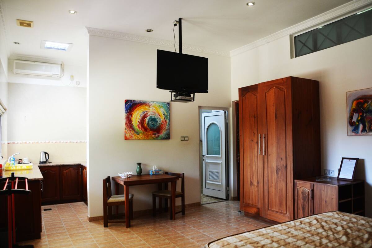 A room with a bed, a wooden wardrobe, a desk with a chair, a television mounted on the wall, and a colorful painting.
