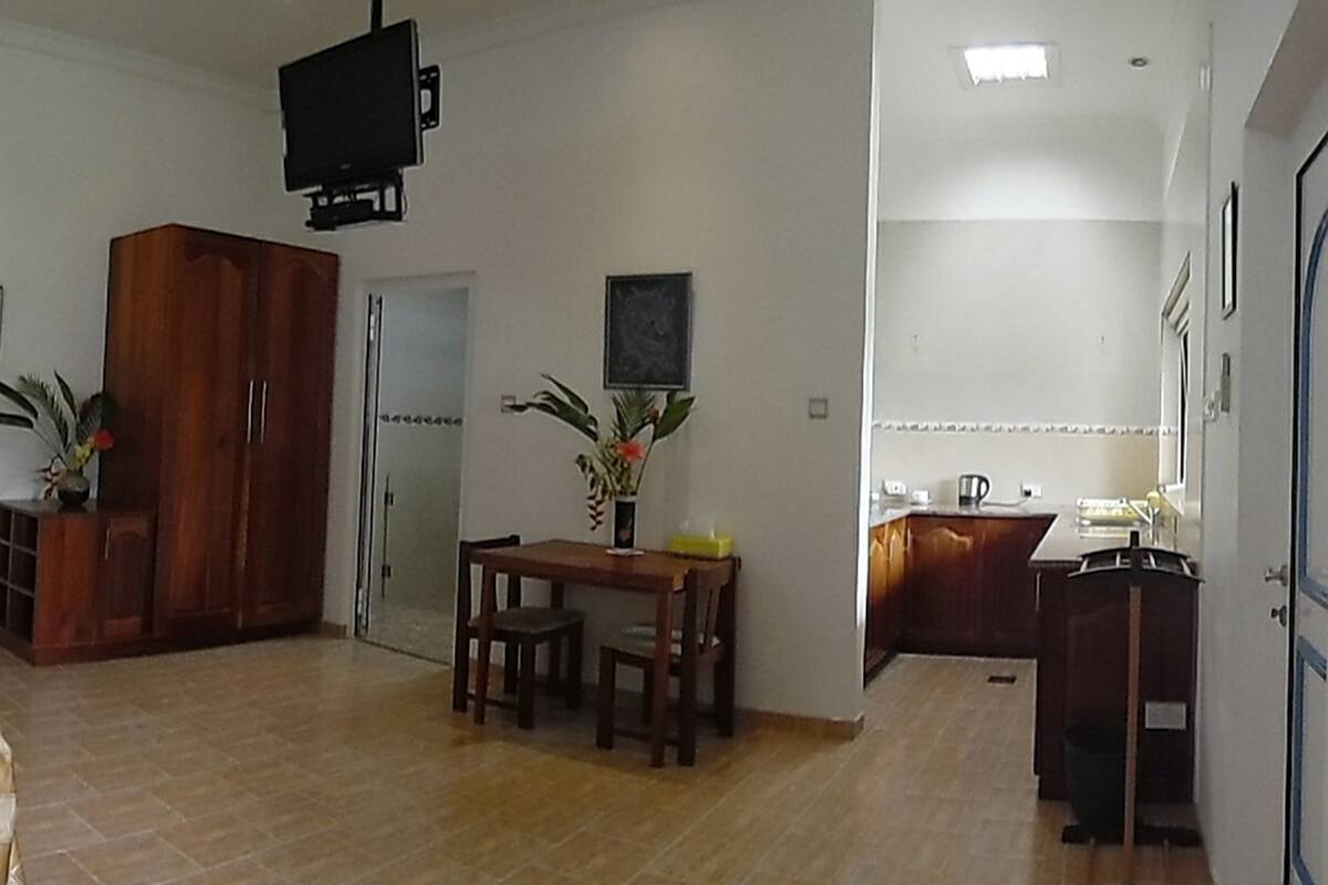 A living room with a wooden cabinet, a dining table with chairs, a TV mounted on the wall, and a kitchen area with a counter and cabinets.