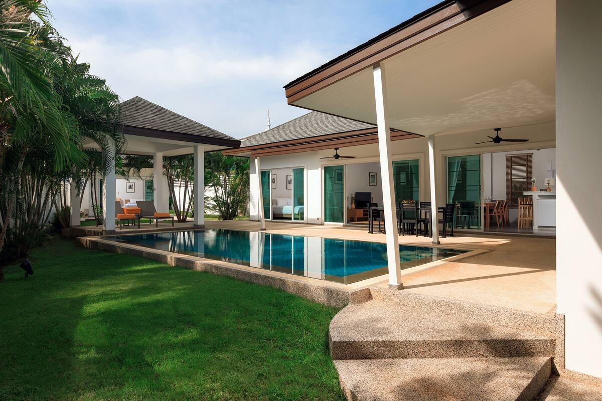 A covered outdoor area with a swimming pool, a patio with seating, and a view of a house in the background.