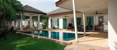 A covered outdoor area with a swimming pool, a patio with seating, and a view of a house in the background.