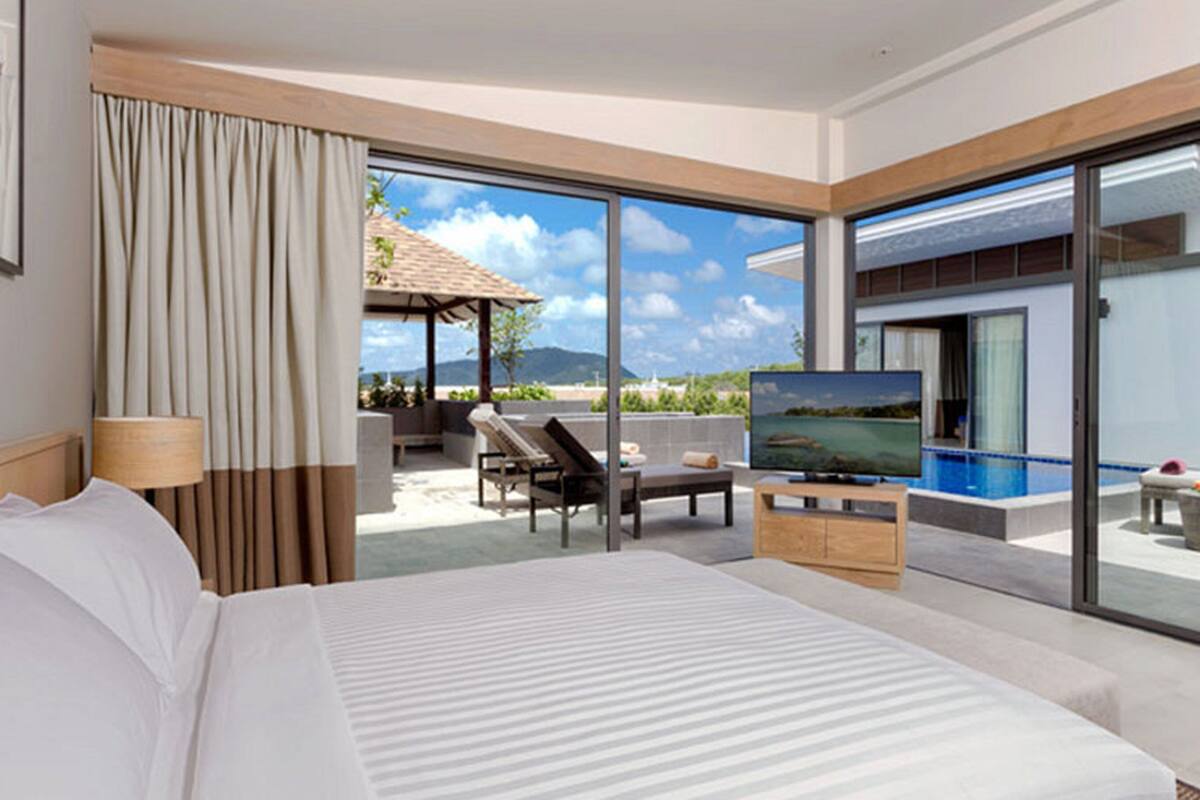 A hotel room with a large bed, a TV, a balcony with a pool, and a view of the mountains.