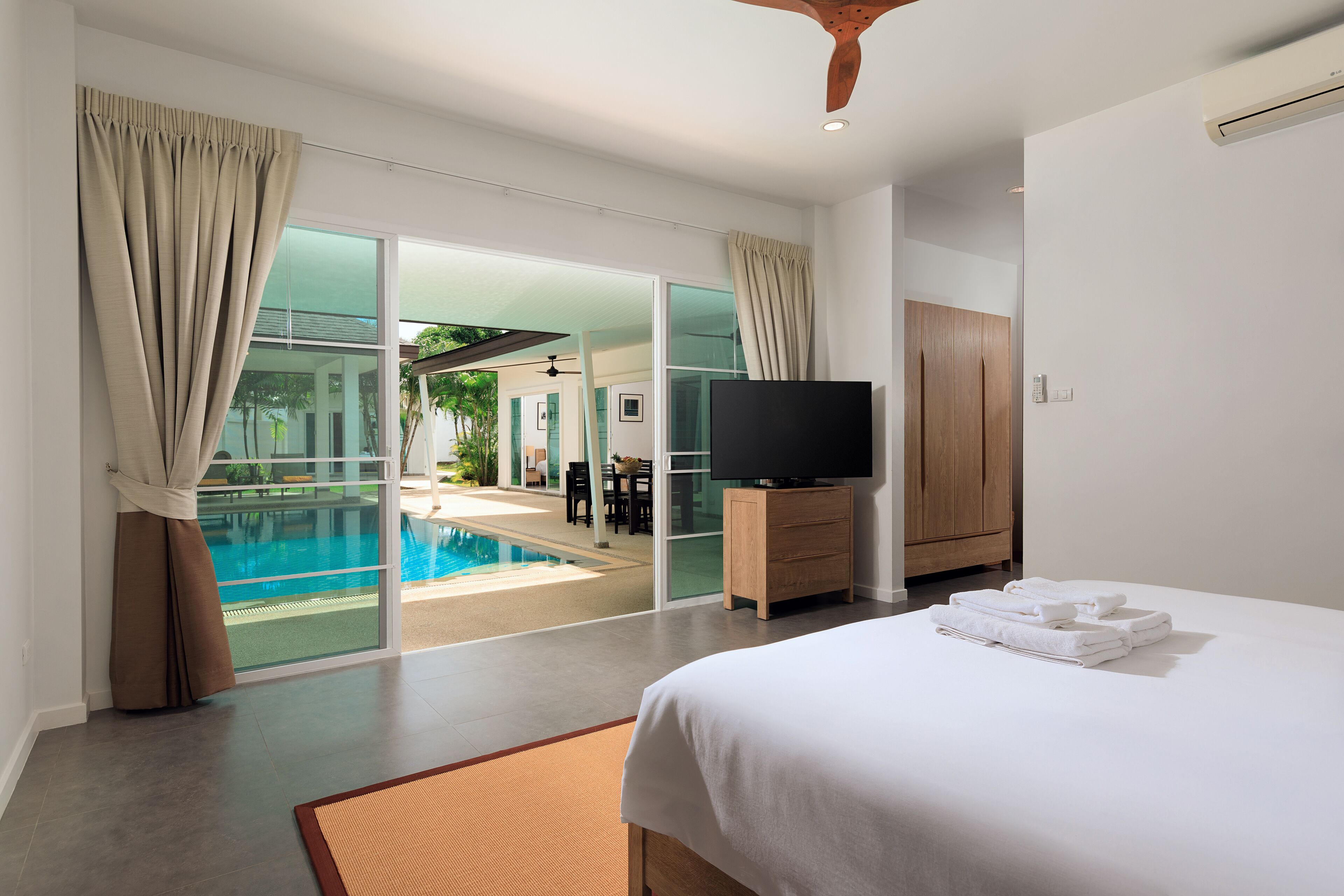 A modern hotel room with a large bed, a flat-screen TV, a wooden cabinet, and a view of an outdoor swimming pool.