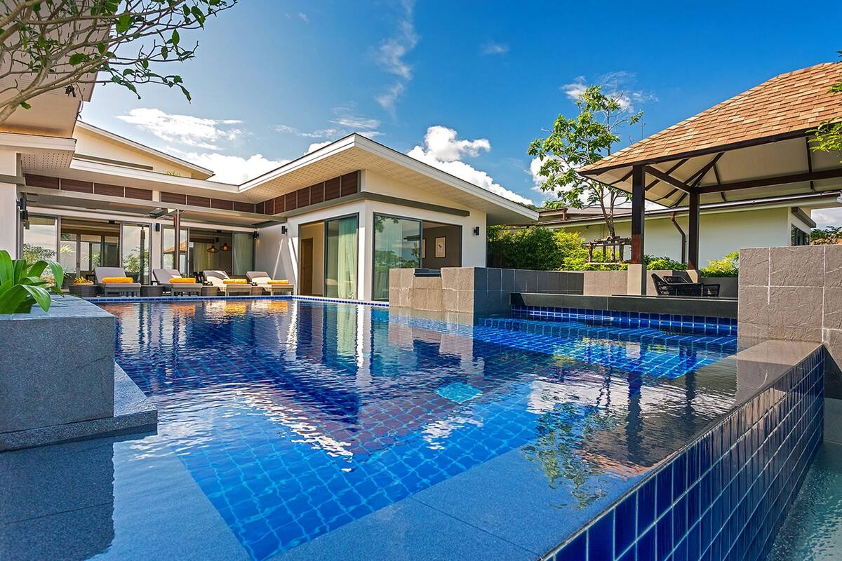 A modern pool area with clear blue tiles, a covered seating area, and a well-maintained garden.