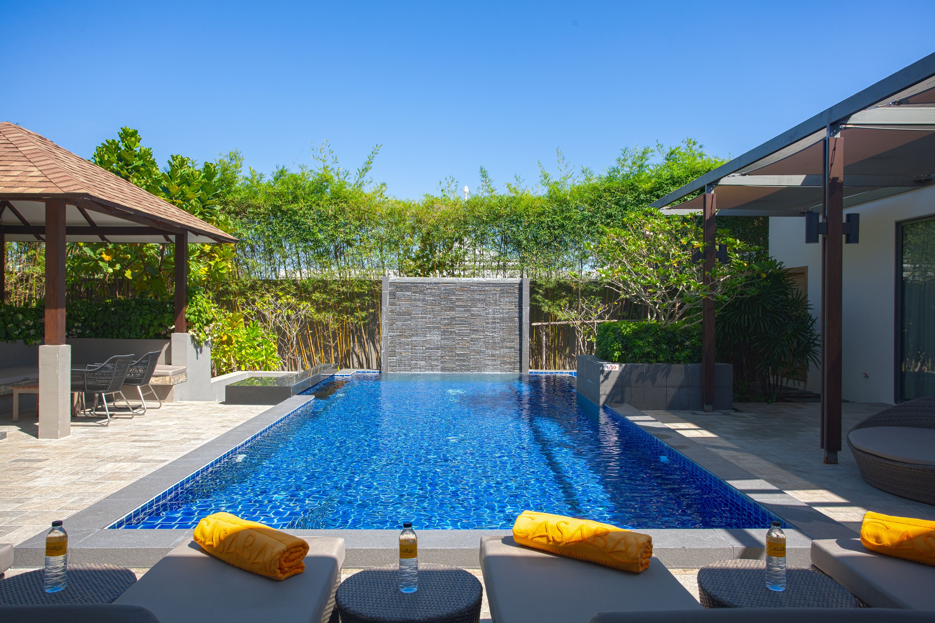 A rectangular swimming pool with a waterfall feature, surrounded by lounge chairs and a pergola.