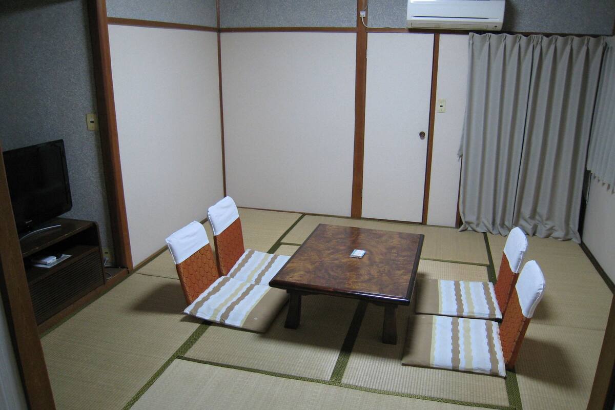 Ein Raum mit Tatami-Matten, einem niedrigen Tisch, Stühlen, einem Fernseher und einer Klimaanlage.