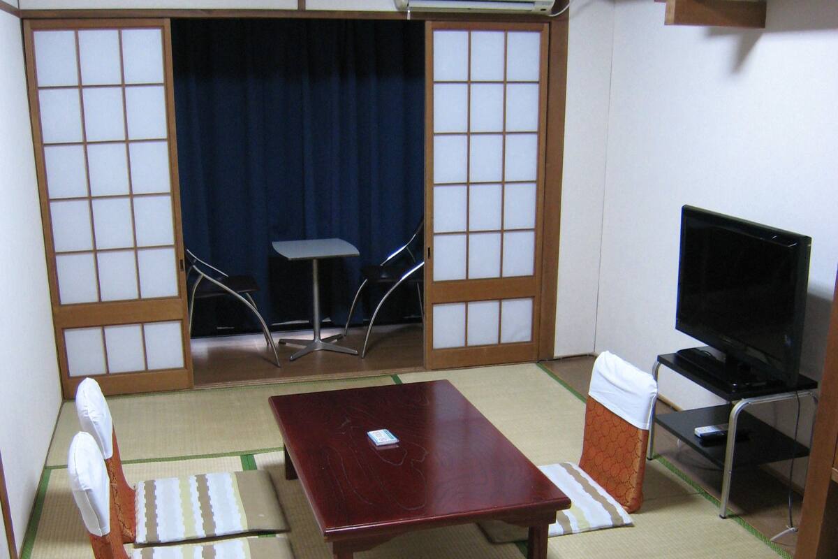Ein traditioneller japanischer Raum mit Tatami-Bodenbelag, einem niedrigen Tisch, einer Couch, einem Fernseher und einer Schiebetür.