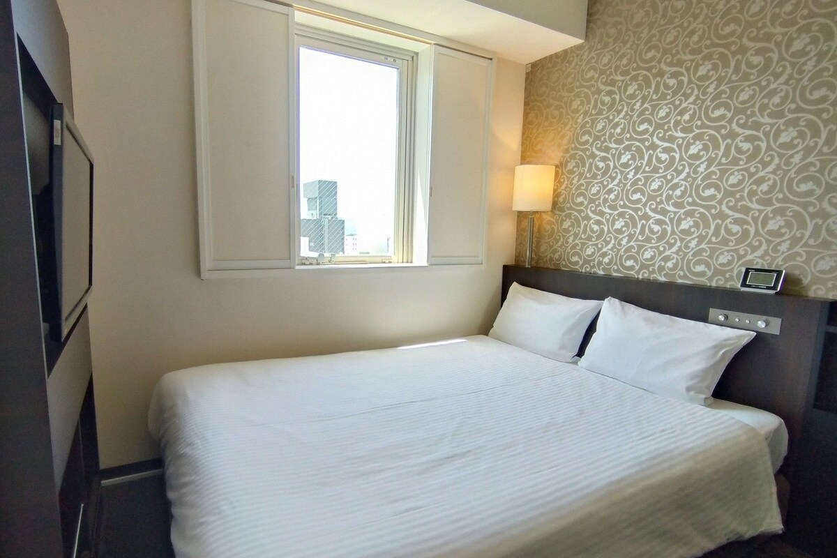 A hotel room with a large bed, a window, a wall clock, and a patterned wallpaper.