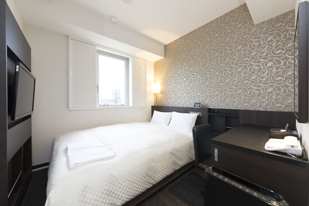 A hotel room with a bed, a desk with a telephone, a TV, and a window.