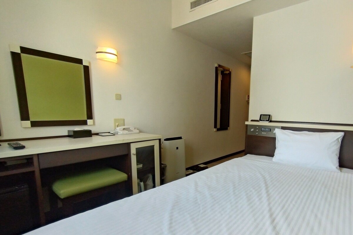 A hotel room with a bed, a desk with a telephone, a mirror, and a wall-mounted light fixture.