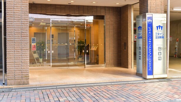 Entrada de hotel con puertas de cristal, un mostrador de recepción y un quiosco informativo azul.