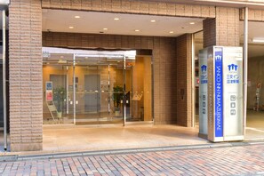 Entrada de hotel con puertas de cristal, un mostrador de recepción y un quiosco informativo azul.