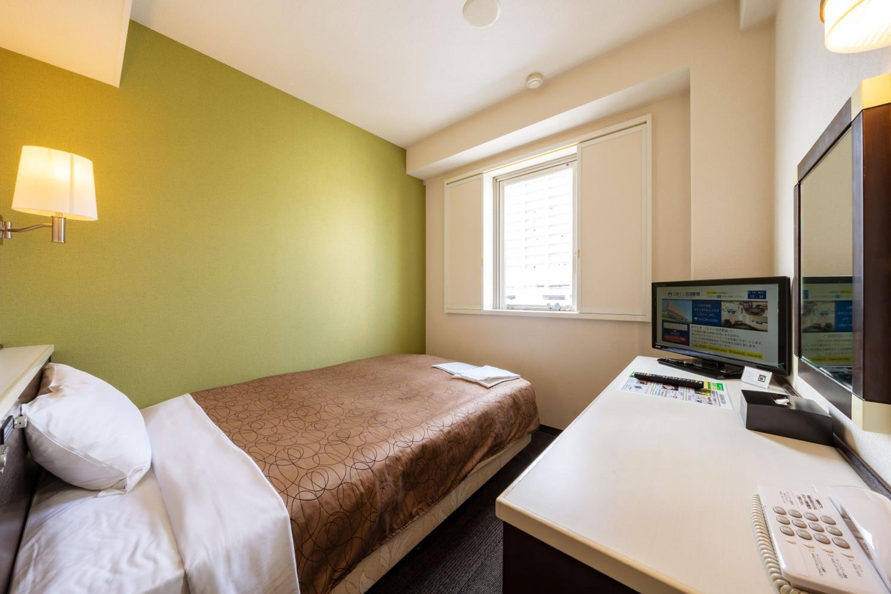 A hotel room with a bed, a desk with a computer, a telephone, and a window with blinds.