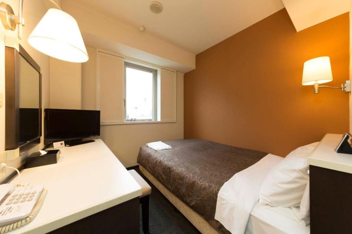 A hotel room with a single bed, a desk with a telephone, a television, and a window with blinds.