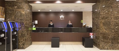 Un comptoir d’accueil avec un mur en marbre, deux réceptionnistes et un kiosque numérique.