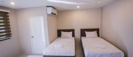 A small, clean room with two single beds, a white door, and an air conditioning unit on the wall.