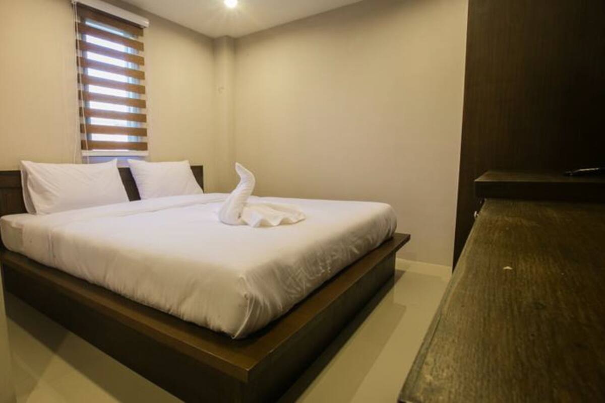 Una cama bien hecha con sábanas blancas y una toalla doblada encima, una ventana con persianas y un cabecero de madera.