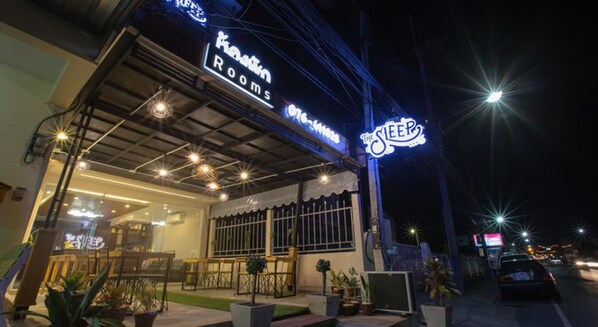 Un hotel llamado "Kavin Rooms" con un letrero de neón que dice "The Sleep" y un área de estar exterior visible con mesas y sillas.