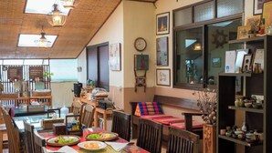 A dining area with a long table set for a meal, surrounded by wooden chairs. There are hanging lights, a clock on the wall, and a shelf with various items. - Gita's House (Chiang Rai)